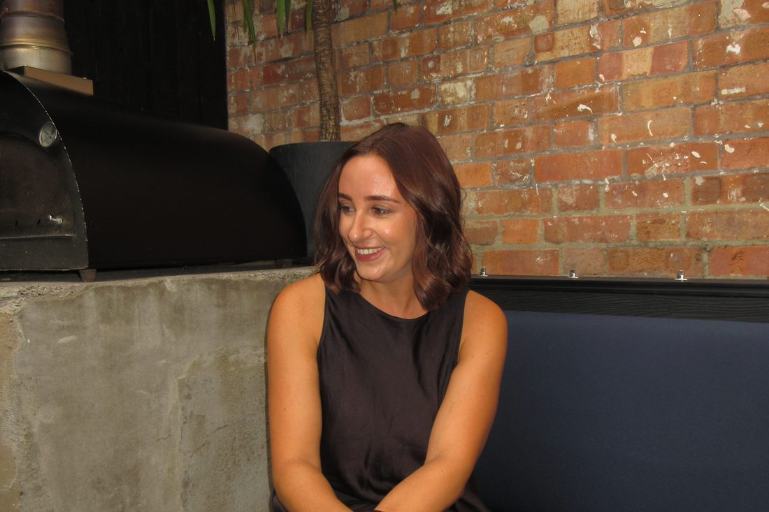 Kate laughing and looking off to one side, seated on a banquette against an exposed brick wall.
