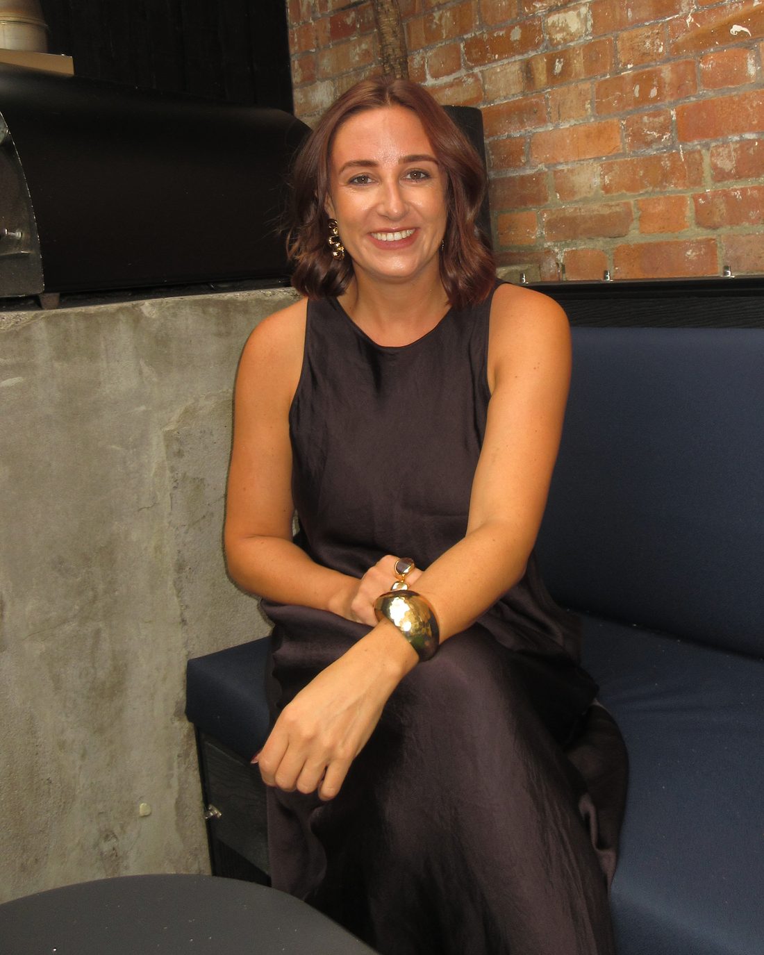 Kate in a black sleeveless dress and gold cuff, smiling at the camera from a navy banquette beside a brick wall.